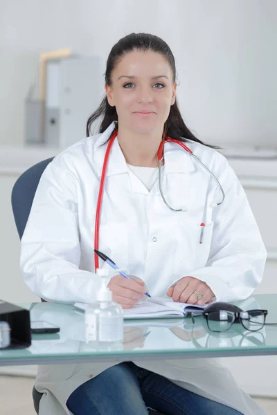 Jovem sorridente feminino médico sentado na mesa e escrevendo — Fotografia de Stock