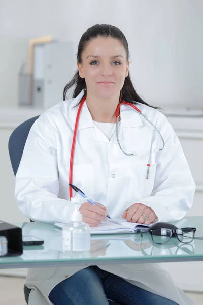 Joven doctora sonriente sentada en el escritorio y escribiendo — Foto de Stock