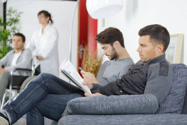 Men sat on settee, patient being wheeled by in wheelchair — Stock Photo, Image