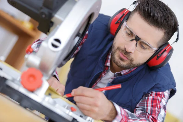 Apuesto carpintero en gafas protectoras que miden tablón — Foto de Stock