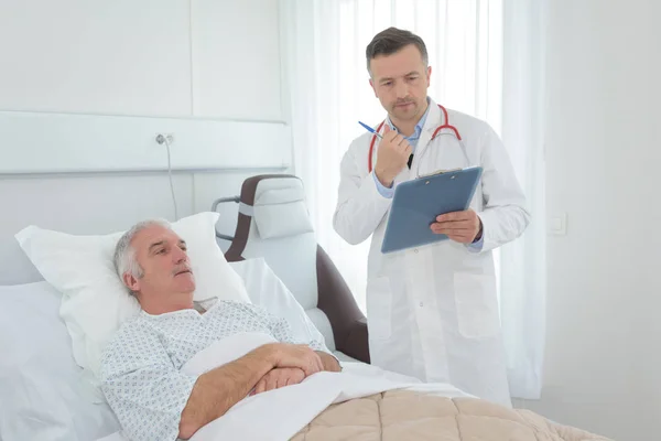 Doctor with clipboard beside patient s bed — Stock Photo, Image