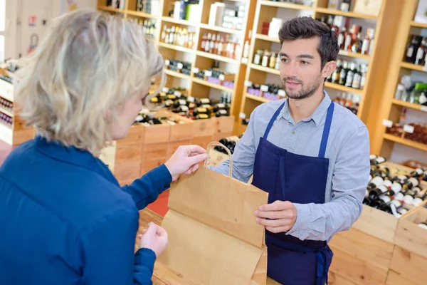 Salesman putting wine bottle in paperbag for customer — Stock Photo, Image