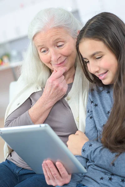 Granddaughter and grandmother with tablet sitting on couch at home — Stock Photo, Image