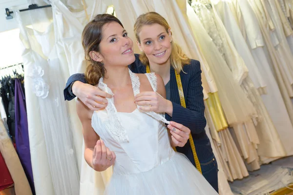 Jovem noiva tentando no vestido de noiva com assistente — Fotografia de Stock