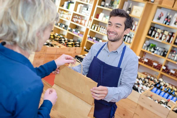 Salesman putting wine bottle in paperbag for customer — Stock Photo, Image