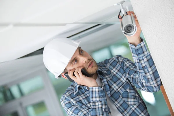 Tecnico fissaggio telecamera cctv a parete e telefono per aiuto — Foto Stock