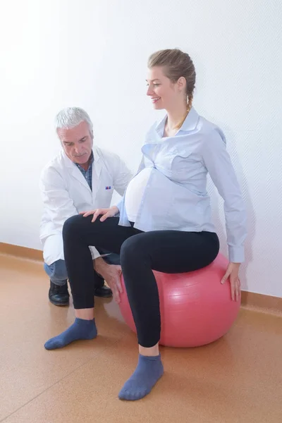 Gravid kvinna sittande på bollen gör excersices med läkare — Stockfoto