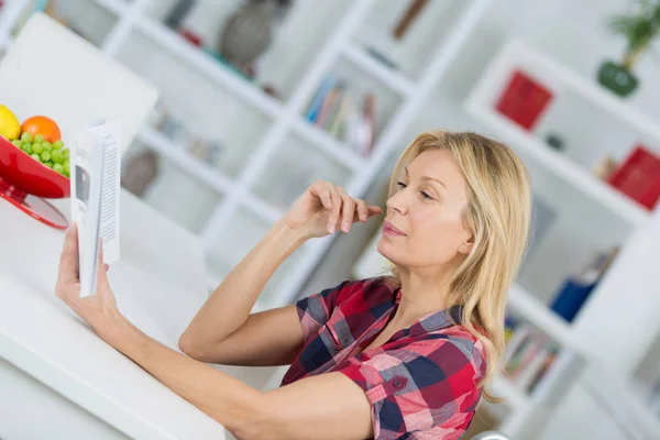 Mulher sorrindo livro de leitura na cozinha em casa — Fotografia de Stock