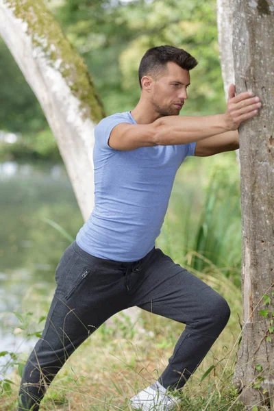 Ung man gör sträcker sig mot ett träd i en park — Stockfoto