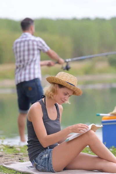 Lady sat reading a book while man fishes — Stock Photo, Image