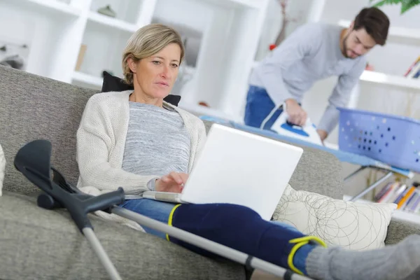 Signora utilizzando il suo computer portatile mentre assistente domestico è stiratura — Foto Stock