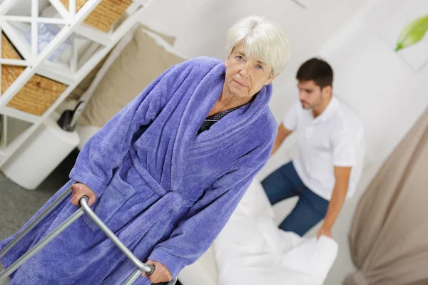 Infirmière mâle faisant lit tandis que la femme aînée utilisant walker — Photo