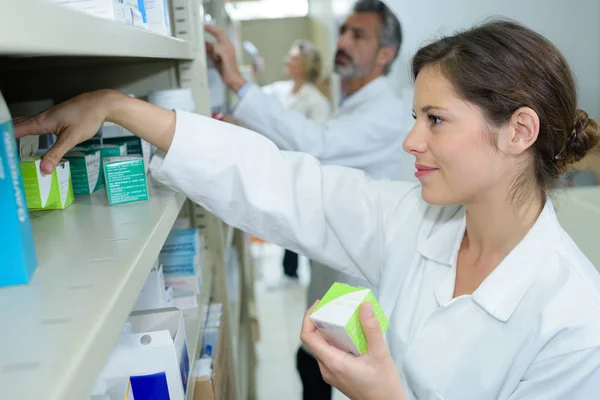 Sonriente joven farmacéutica almacenamiento de medicamentos en estantería — Foto de Stock