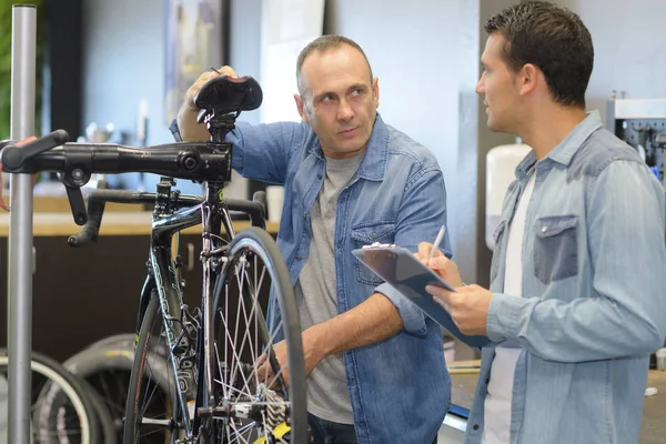 Vendedor em uniforme falando com o cliente durante a fixação de bicicleta — Fotografia de Stock