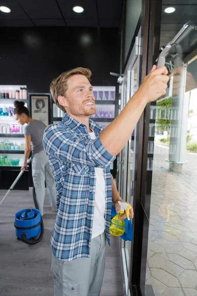 Young cleaner wiping a window at hairdresser salon — Stock Photo, Image