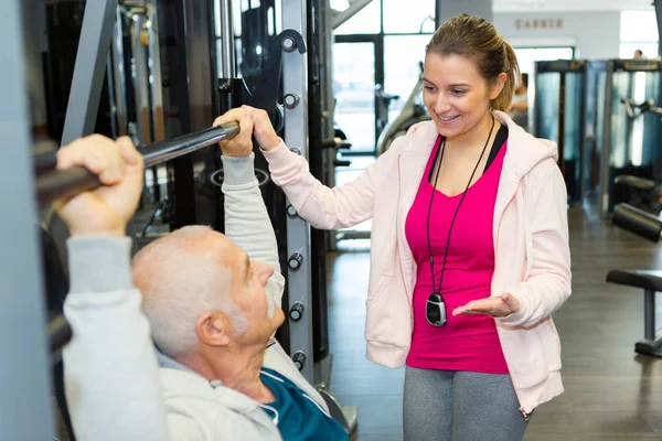 Entrenador femenino que ayuda al hombre mayor a levantar peso en el gimnasio — Foto de Stock
