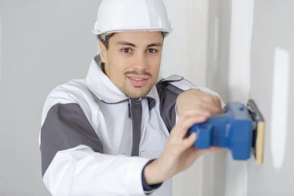 Jovem construtor em hardhat lixar parede dentro de casa — Fotografia de Stock