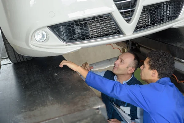 Mecánico y masculino aprendiz trabajando debajo del coche juntos — Foto de Stock