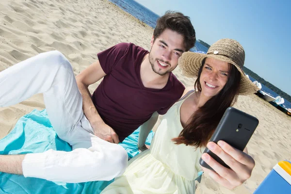 Великолепная молодая пара, сидящая на пляже и делающая селфи — стоковое фото