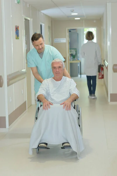 Enfermera tomando hombre mayor paciente en silla de ruedas en el pasillo del hospital —  Fotos de Stock