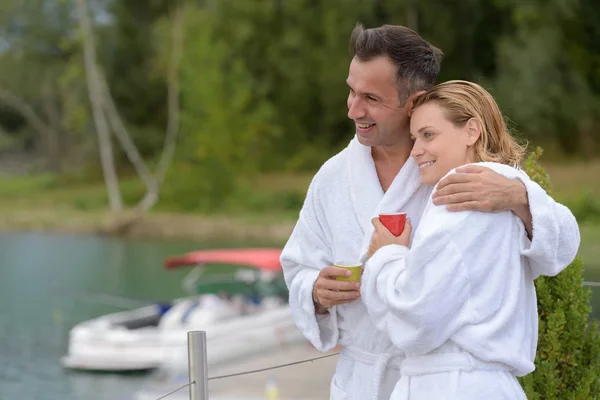 Embracing couple in bathrobes looking out over water — Stock Photo, Image