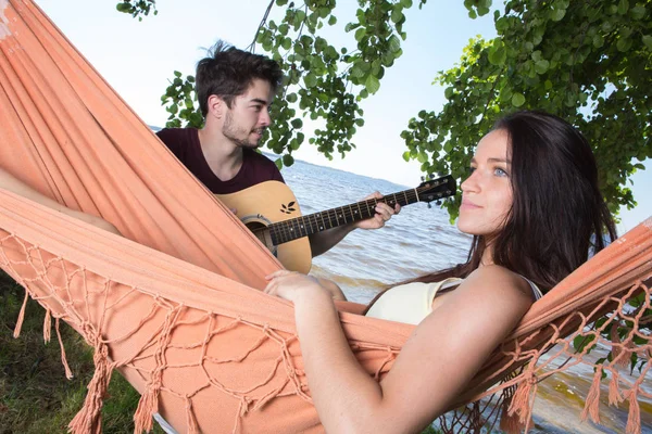 Menina bonita relaxante em rede ouvindo seu namorado tocando guitarra — Fotografia de Stock