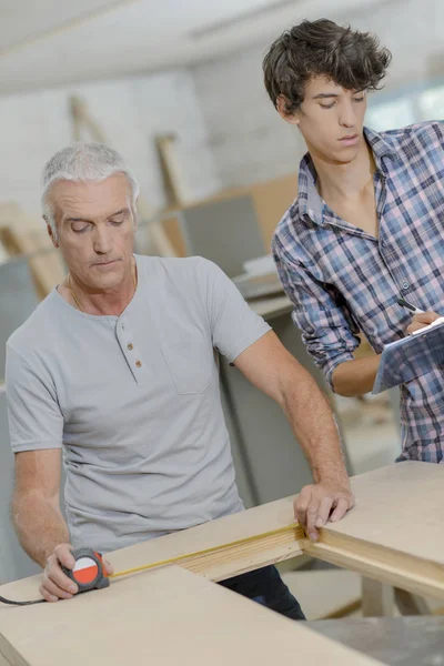 Vedoucí carpenter měřící otvor do dřeva, junior psaní poznámek — Stock fotografie