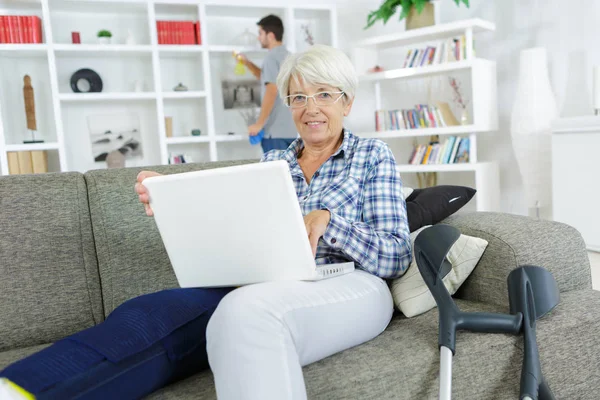 Senior woman with laptop and injured leg at home — Stock Photo, Image