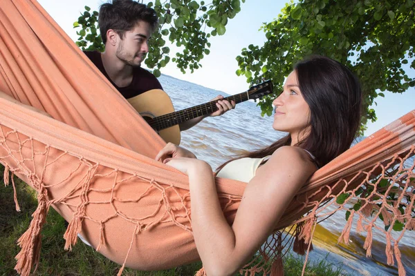 Menina bonita relaxante em rede ouvindo seu namorado tocando guitarra — Fotografia de Stock