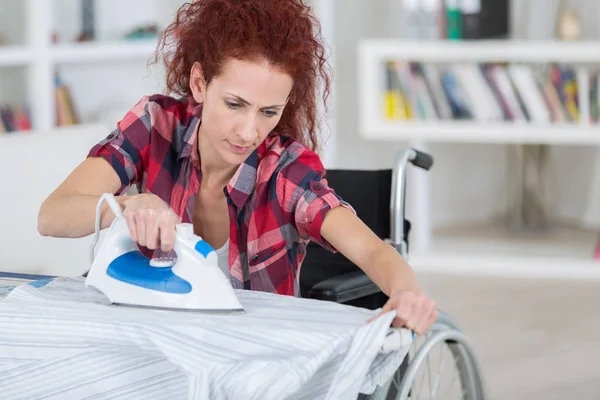 Pretty redhead woman sitting on wheelchair and ironing — Stock Photo, Image