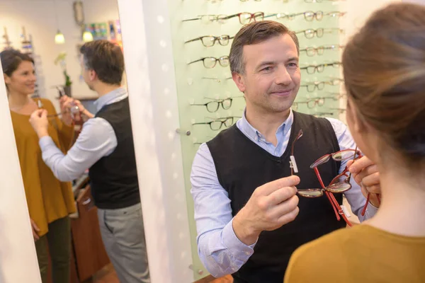 Oftalmóloga y hermosa morena hembra eligiendo gafas en tienda de óptica —  Fotos de Stock