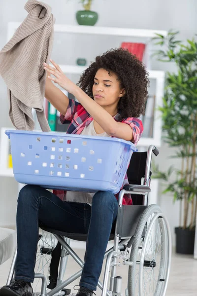 Ung glad kvinna på rullstolen sätta kläder i korg — Stockfoto