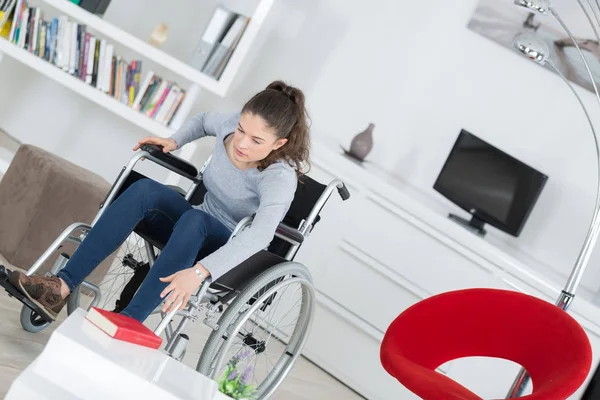 Mujer discapacitada tratando de conseguir libro silla de ruedas en la sala de estar — Foto de Stock