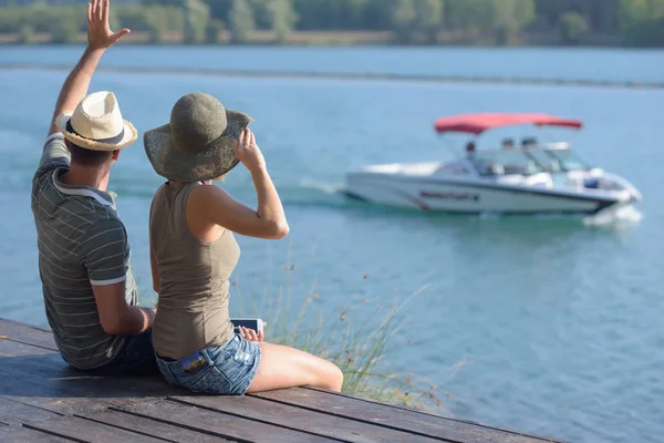 Paar saß auf Seebrücke und winkte vorbeifahrendem Boot zu — Stockfoto