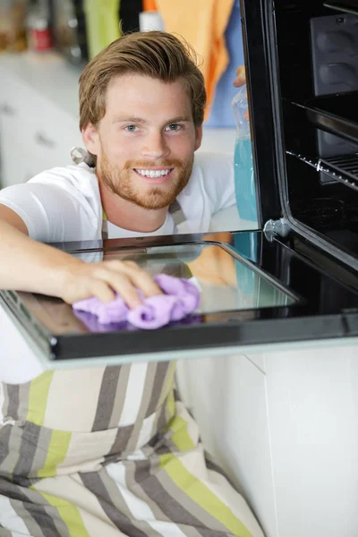 Happy young man in overall cleaning oven with rag — Stock Photo, Image