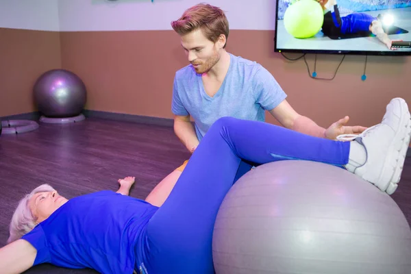 Mujer mayor en el gimnasio haciendo ejercicio en fitball con el entrenador — Foto de Stock