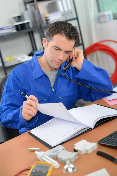 Mann in Arbeitskleidung am Telefon, Blick auf Tagebuch — Stockfoto