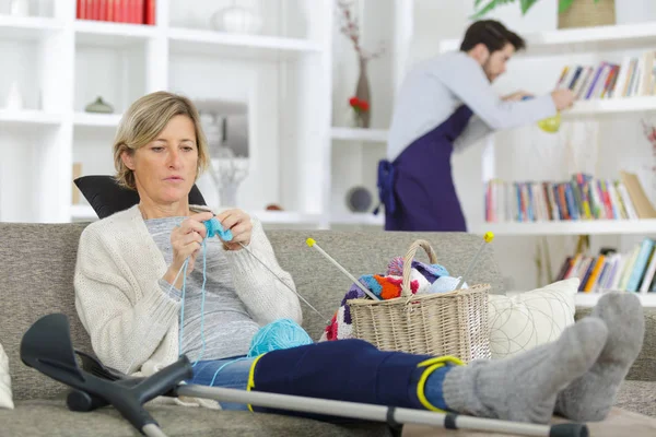 Injured lady knitting while man dusts shelves in background — Stock Photo, Image