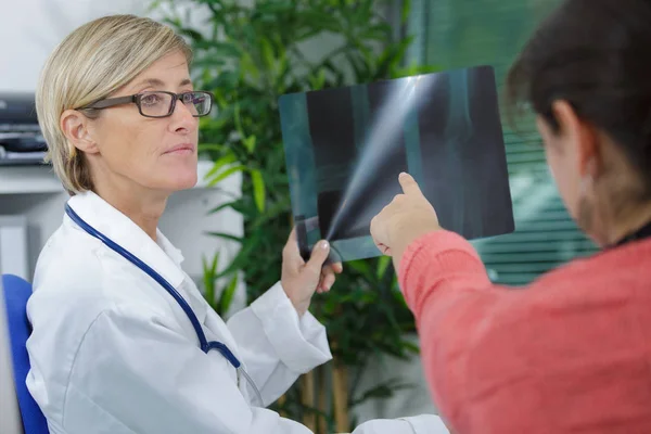 Médico femenino sosteniendo una película de rayos X mientras el paciente señala —  Fotos de Stock