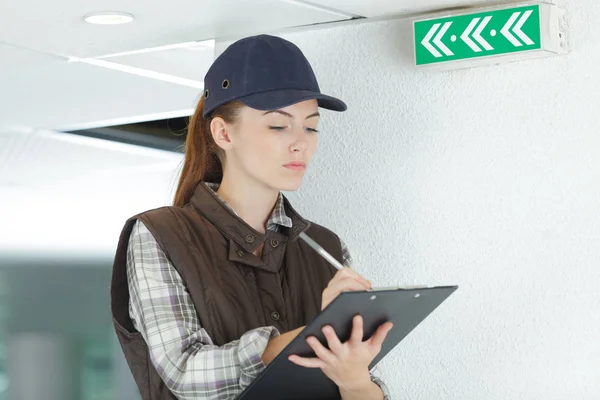 Female inspector with clipboard next to green exit sign — Stock Photo, Image