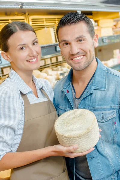 Shop assistant holding cheese, stood next to customer — Stock Photo, Image
