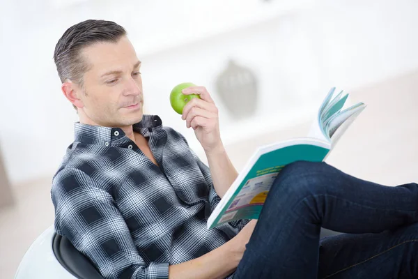 Man eating apple and reading while laying on the couch — Stock Photo, Image