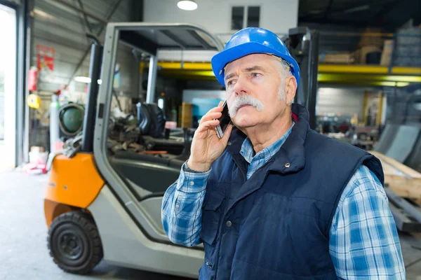 Старший работник на вилочном погрузчике говорит по мобильному телефону — стоковое фото