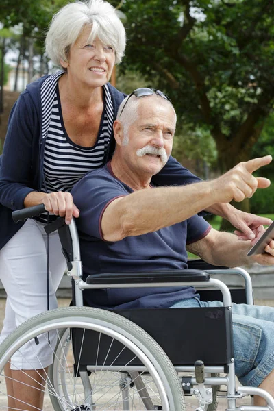Пожилая женщина толкает своего мужа на инвалидной коляске в парке — стоковое фото