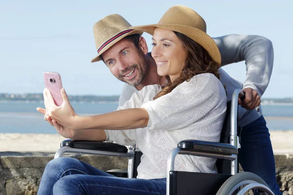 Couple au bord de la plage en fauteuil roulant prenant selfie — Photo