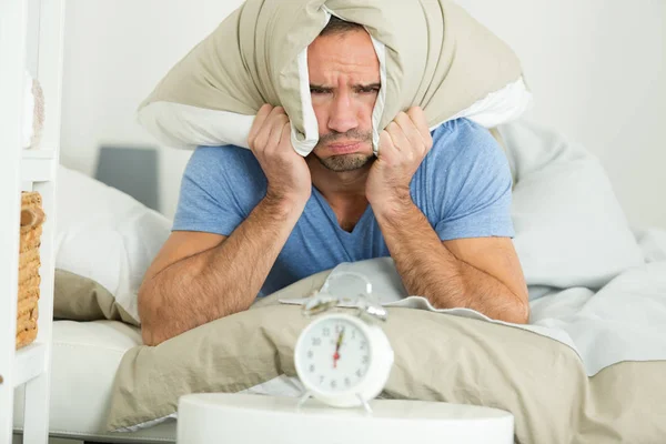 Man covering his ears with pillow looking at alarm clock — Stockfoto