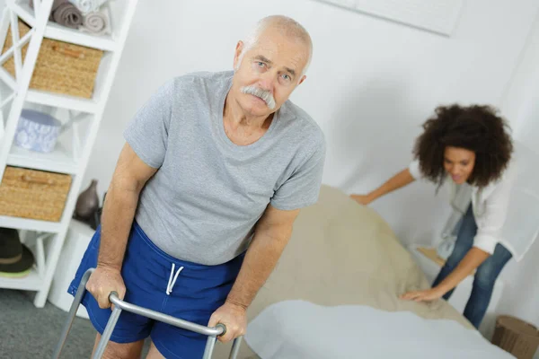 Senior Man Walking Frame Carer Ścielenie Łóżka Tle — Zdjęcie stockowe