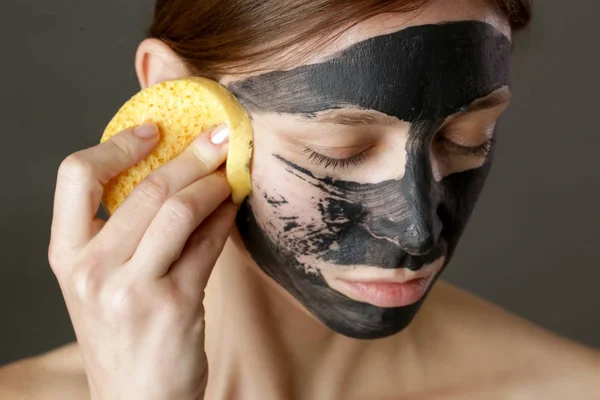 Charcoal face mask or black clay mud. Beautiful woman removing black mask from her face, close up studio head shot. Natural cosmetic concept.