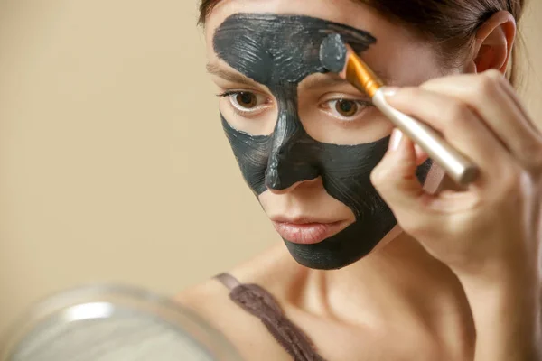 Charcoal face mask or black clay mud. Beautiful woman is applying purifying black mask on her face, close up studio shot. Home spa natural cosmetic concept.