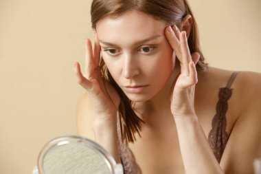Portrait of a young brunette woman, in her thirties, examining her face in front of mirror, aging, fine wrinkles and skin complexion concept clipart
